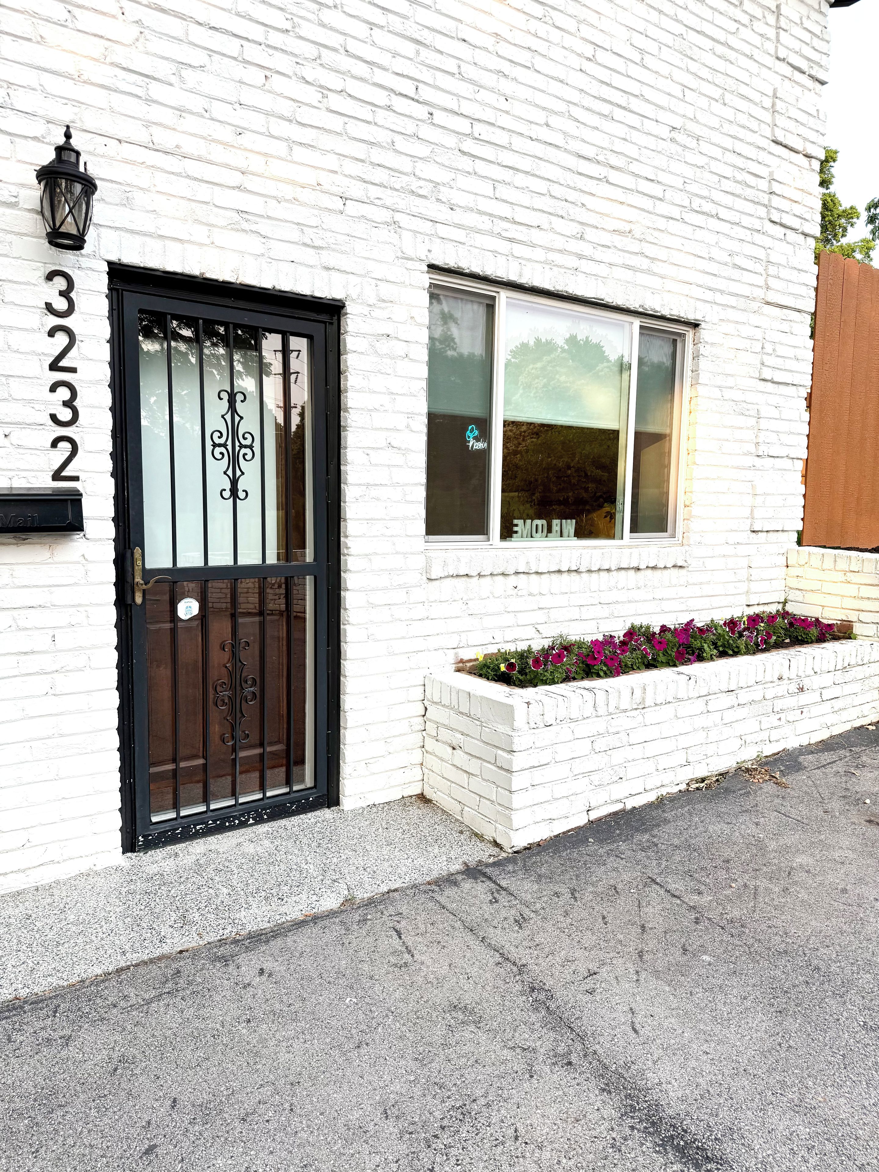A white brick building with a black metal gate, a window with a frosted glass, and a flower bed with purple flowers.