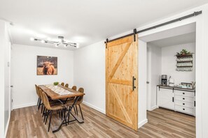 A dining area with a wooden table and chairs, a sliding barn door, a kitchen with white cabinets, and a ceiling light fixture.