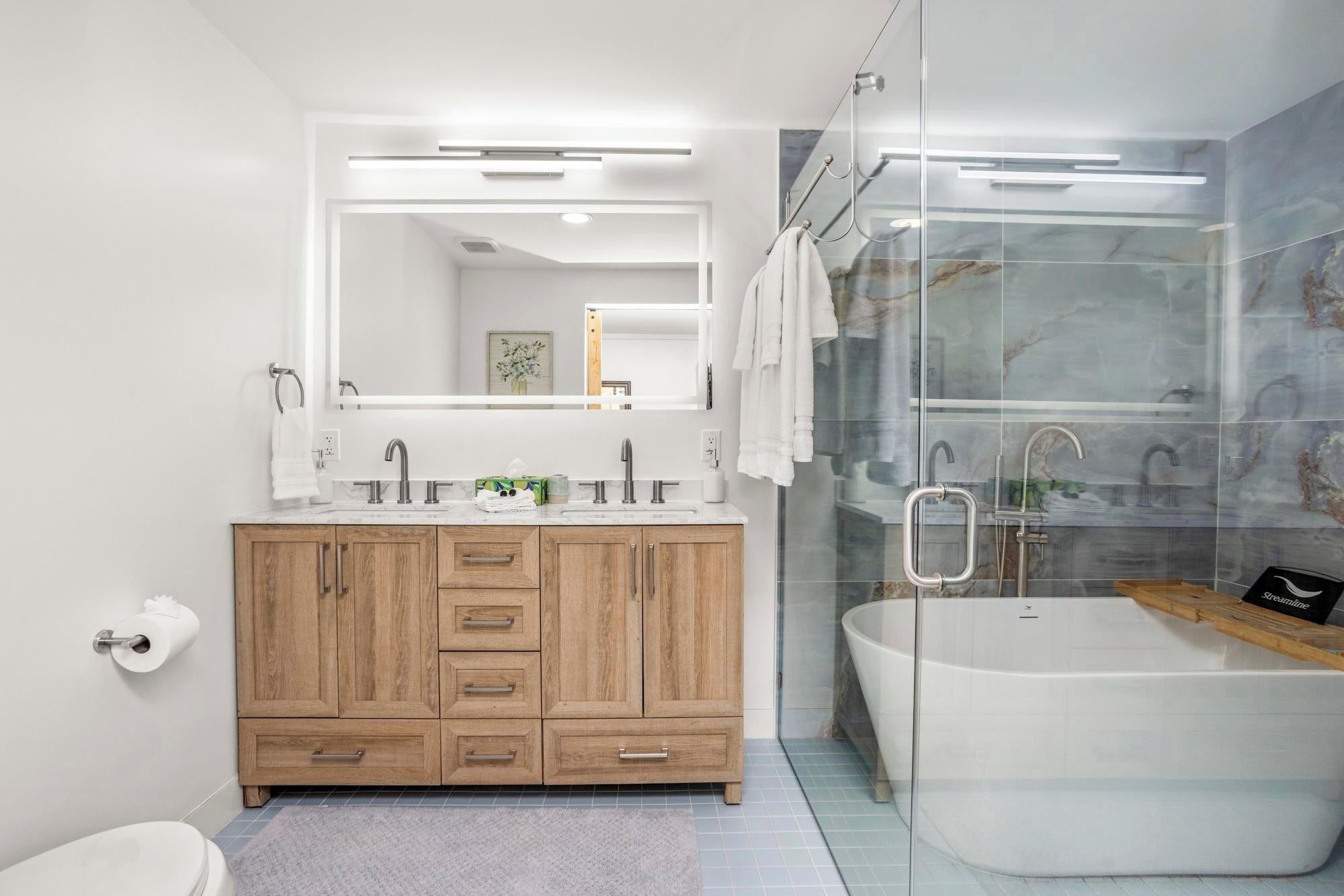 A modern bathroom with a wooden vanity, a large mirror, a glass shower enclosure, and a freestanding bathtub.