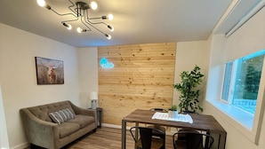 Una sala de estar acogedora con un sofá, una mesa de comedor de madera con sillas, una pared de madera decorativa, una lámpara de techo y una ventana con vistas.