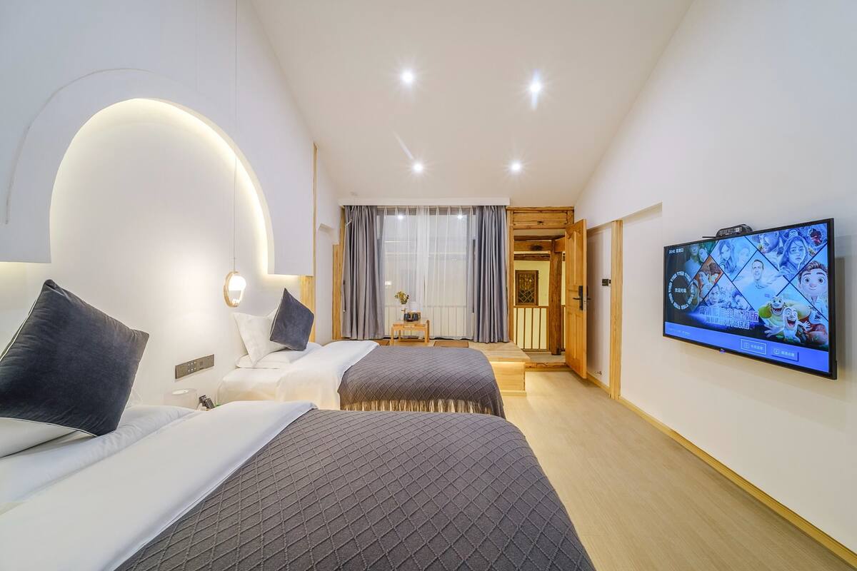 A modern hotel room with two beds, a TV on the wall, and a view of the outside through a large window.