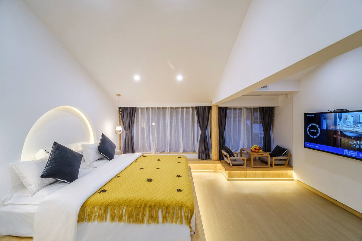 A modern hotel room with a large bed, a TV on the wall, and a seating area with a table and chairs.