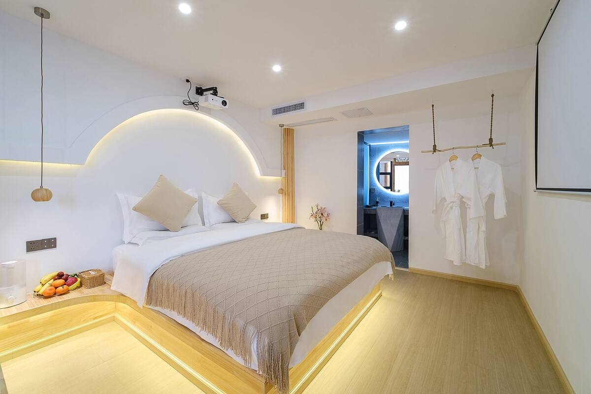 A modern hotel room with a large bed, bedside table, and a small seating area. There is a wall-mounted projector and a mirror with a circular frame.