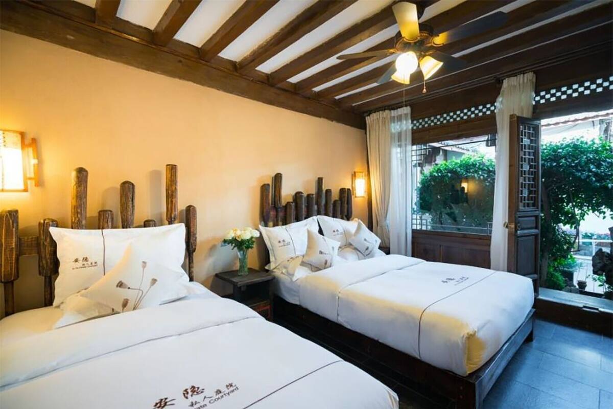 A hotel room with two beds, wooden headboards, a ceiling fan, and a view of greenery outside.