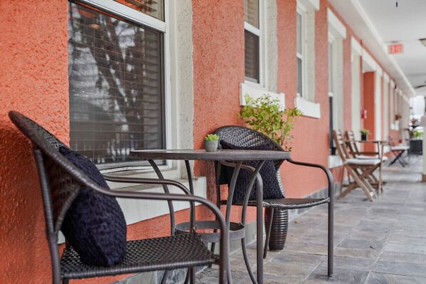 A row of outdoor tables and chairs with wicker furniture, a potted plant, and a building with a textured facade.
