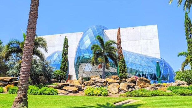 Un bâtiment moderne doté d’une structure en forme de dôme de verre, entouré de palmiers et de rochers.