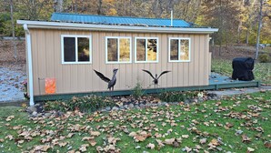 A small, single-story building with a blue roof and beige walls, surrounded by a grassy area with scattered fallen leaves. - The Riverside Cottage... Charming  2-bedroom cottage on the Mohican River (Loudonville)
