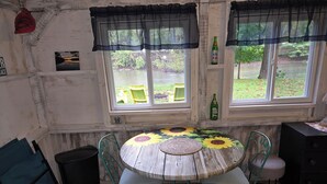 A rustic room with a round table adorned with sunflowers, two windows with sheer curtains, and a bottle of 7UP on the windowsill. - The Riverside Cottage... Charming  2-bedroom cottage on the Mohican River (Loudonville)