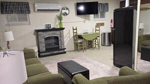 A living room with green upholstered furniture, a fireplace, a television, and a kitchen area with a refrigerator and a sink. - The Riverside Cottage... Charming  2-bedroom cottage on the Mohican River (Loudonville)