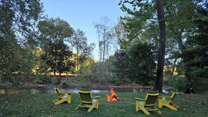 A campsite with three green chairs arranged in a semi-circle around a campfire by a river, surrounded by trees and grass. - The Riverside Cottage... Charming  2-bedroom cottage on the Mohican River (Loudonville)