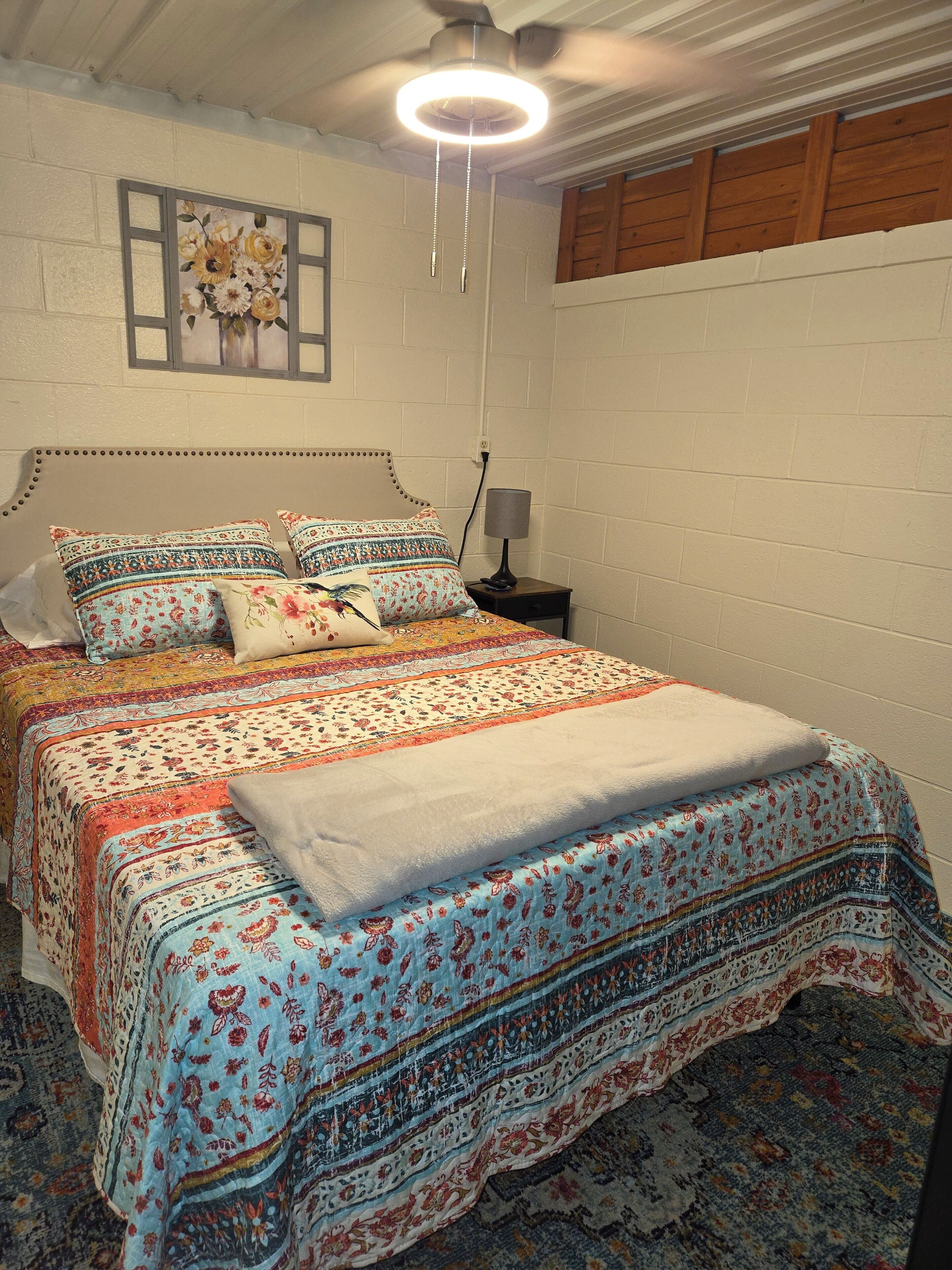 Une chambre à coucher comprenant un lit, une table de chevet, une lampe, un ventilateur de plafond et une œuvre d’art florale accrochée au mur.
