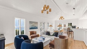 A spacious living area with a white ceiling, large windows, and a kitchen area visible through an open door.