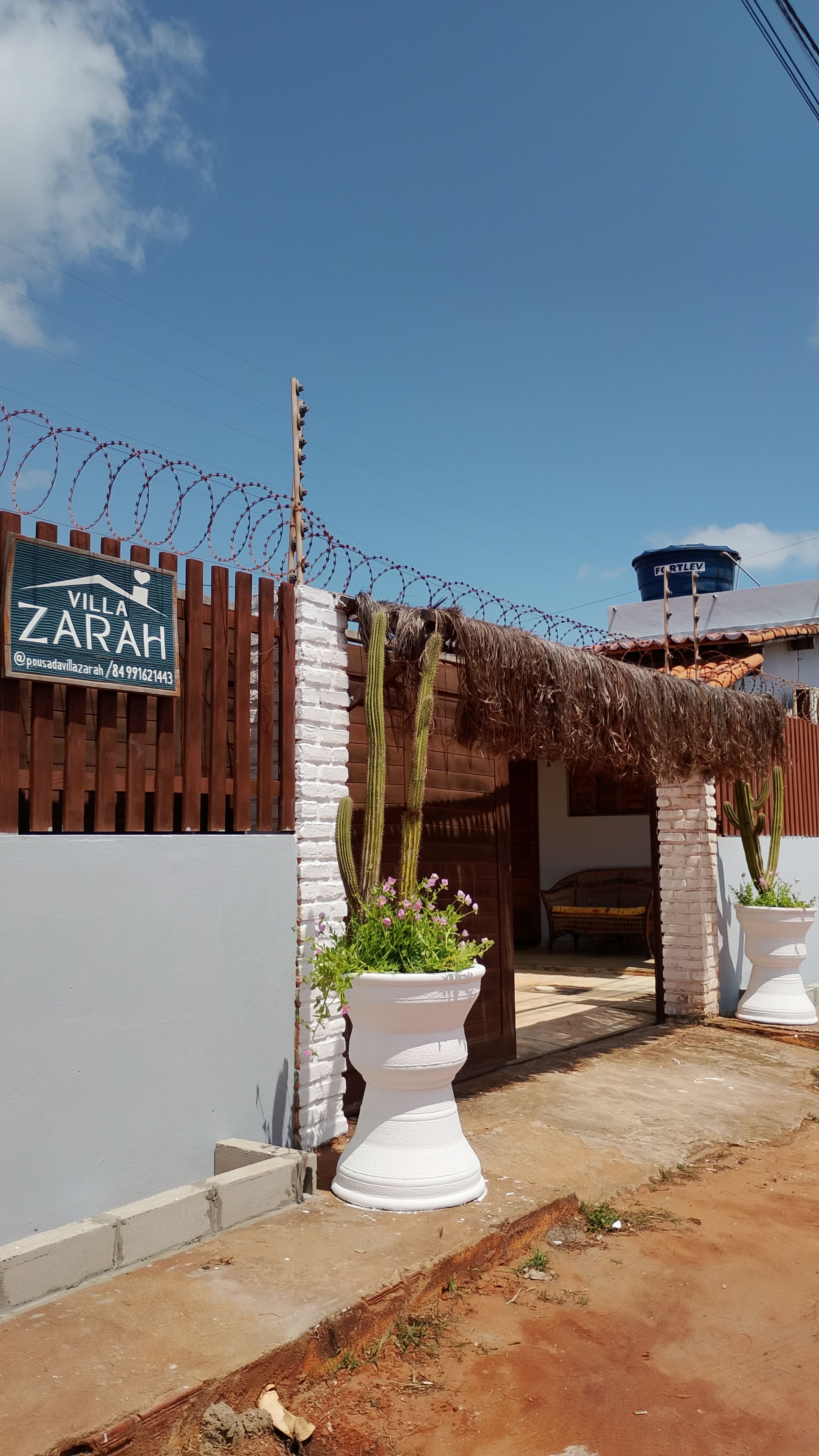 Entrada gradeada com placa indicando 'Villa Zarah', jardim de cactos e um reservatório de água.