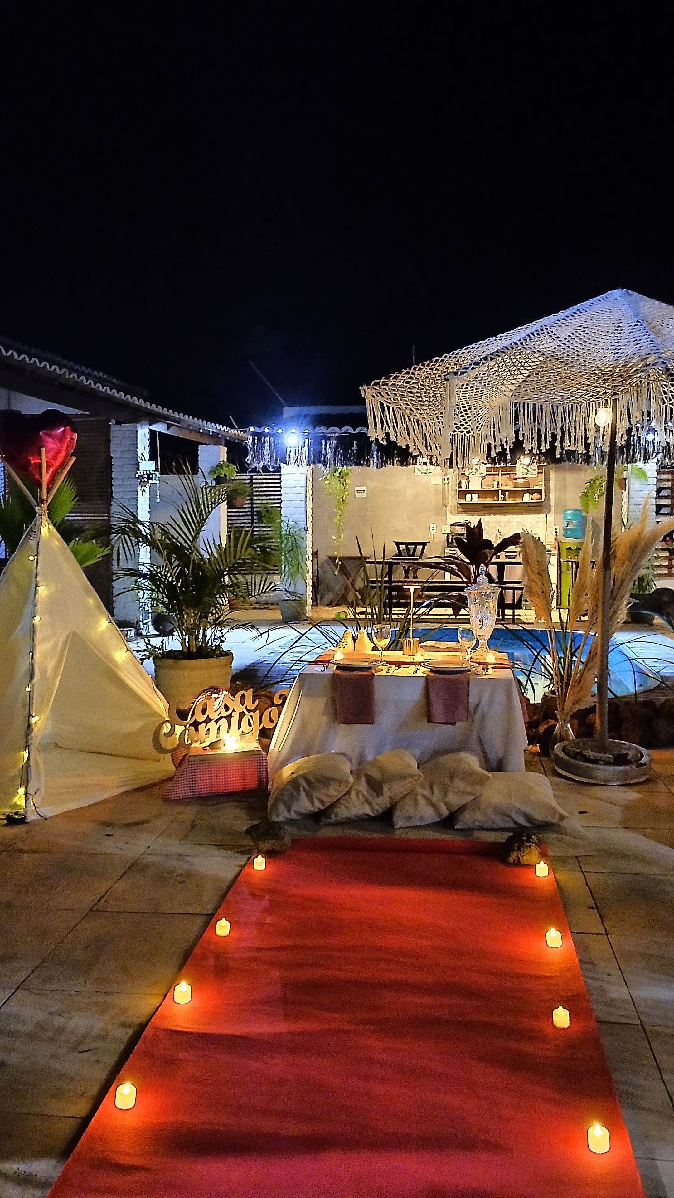 Uma área de refeições ao ar livre com tapete vermelho, mesas montadas com velas e uma estrutura de toldo.