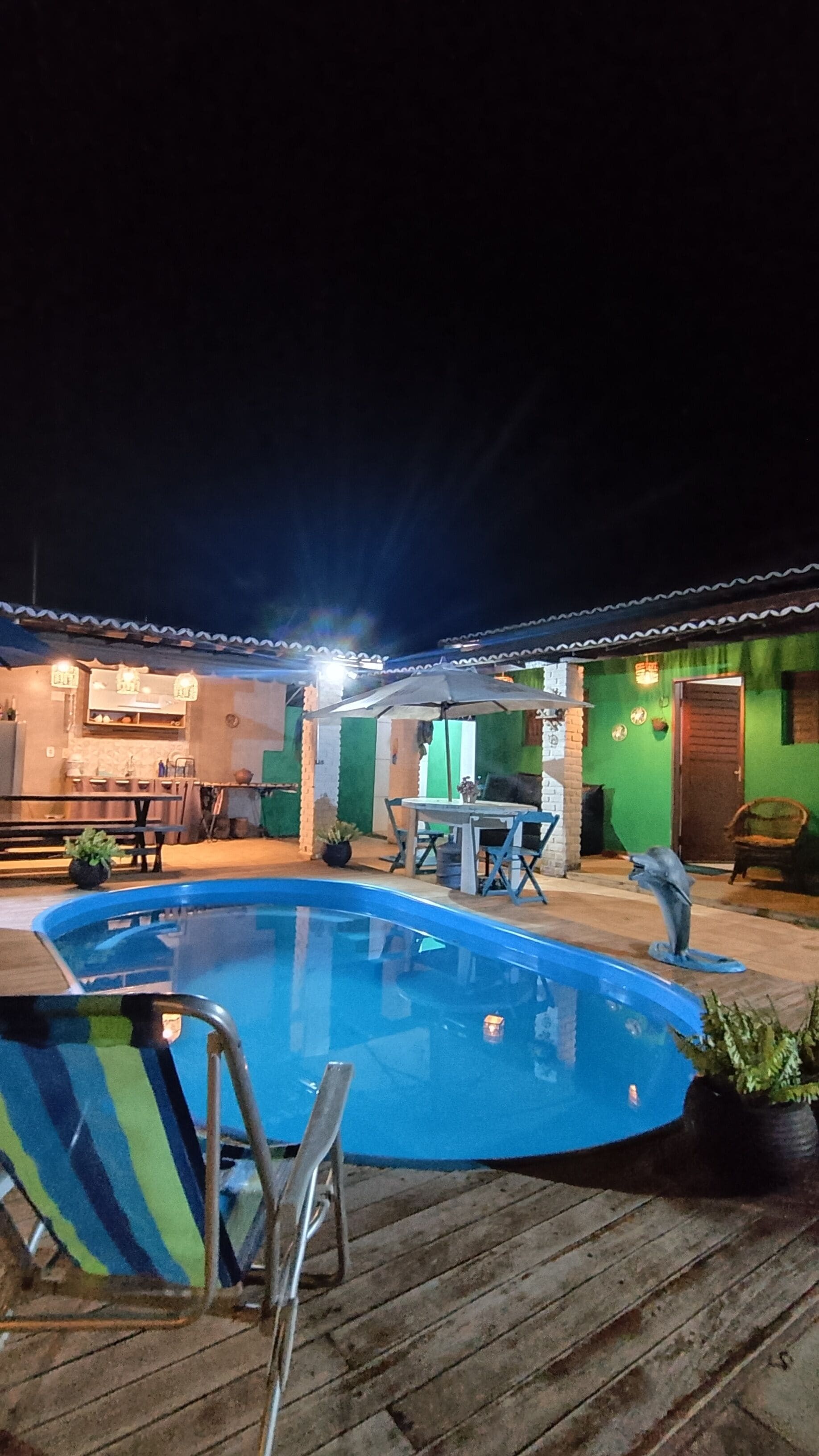 A swimming pool with a wooden deck, a table with chairs, and a building with a green facade in the background.