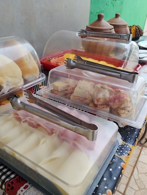 A food display with various items covered in plastic, including what appears to be bread, pastries, and possibly some meat. - Pousada Villa Zarah (São Miguel do Gostoso)