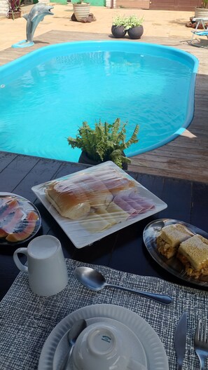 A dining table with a meal, a pool, and a dolphin statue. - Pousada Villa Zarah (São Miguel do Gostoso)