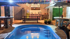 Área externa com piscina, mesa e cadeiras, uma fonte de água e um painel de fundo verde.