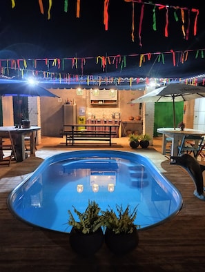 Zona de piscina al aire libre con una mesa y sillas para comer, una fuente de agua y un telón de fondo verde.