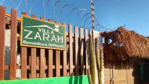 Placa de 'Villa Zarah' com cerca de madeira e arame farpado, um cacto em vaso branco e uma estrutura com telhado de palha.