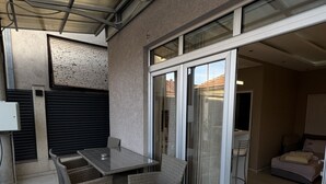 A covered patio area with a table and chairs, a sliding glass door leading to a bedroom, and a black mat at the entrance.