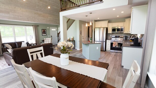 Amplia sala de estar con una mesa de comedor, sillas blancas y un jarrón de flores. Una escalera conduce a un nivel superior.