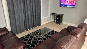 A living room with a black and white patterned rug, a brown leather sofa, a black desk with drawers, and a television displaying 'NETHERLANDS'.