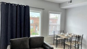 A modern living room with a grey sofa, a dining table with chairs, and large windows with curtains.