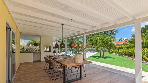 A covered outdoor dining area with a wooden table and chairs, hanging pendant lights, and a view of a swimming pool and greenery. - Kas di Rosa, 8p luxury villa, private pool, large outdoor area (Grote Berg)