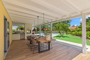 A covered outdoor dining area with a wooden table and chairs, hanging pendant lights, and a view of a swimming pool and greenery. - Kas di Rosa, 8p luxury villa, private pool, large outdoor area (Grote Berg)