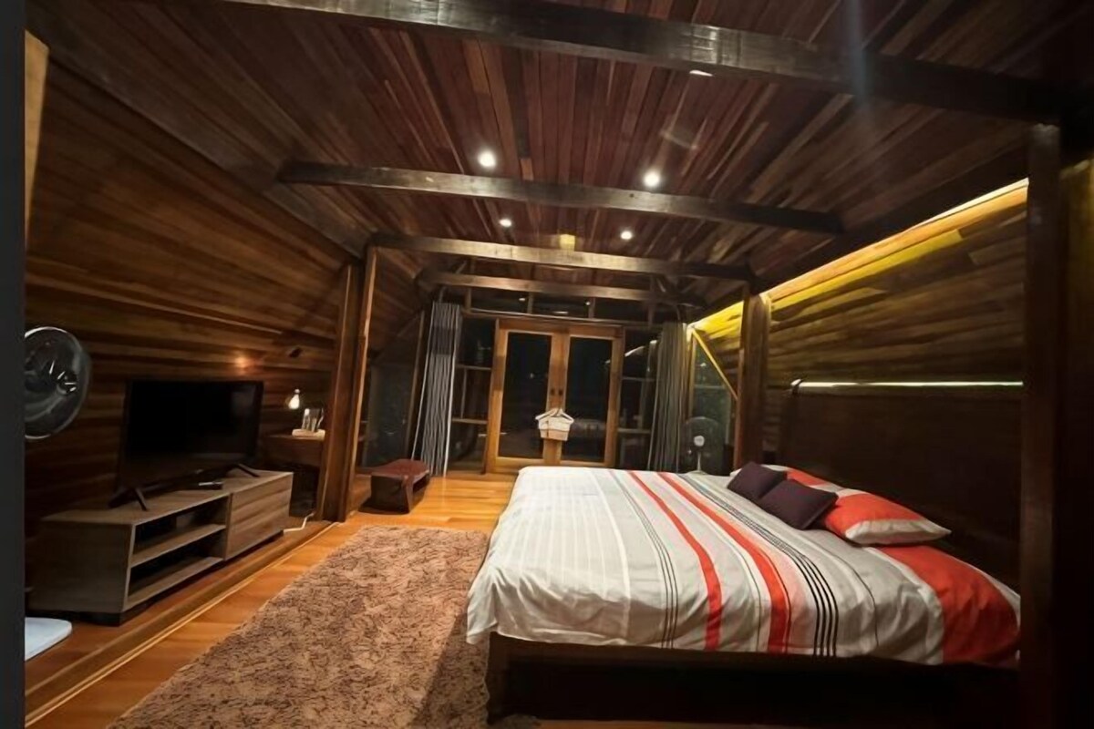 A bedroom with a wooden ceiling, a bed with striped bedding, a TV, and a small table.