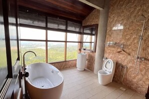 Un baño con bañera, inodoro y ducha, ventanas amplias con vistas, y techo de madera.