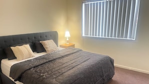 A bedroom with a dark-colored bed, a nightstand with a lamp, and a window with blinds. - Quiet Abode with convenience. 3 Large bedrooms, whole house. (Melton South)
