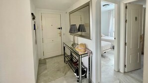 Un pasillo con una puerta blanca, un espejo y una mesita con una lámpara.