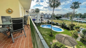 Un balcón con una mesa y sillas de vidrio, una piscina pequeña y vistas a una zona residencial con palmeras y edificios.
