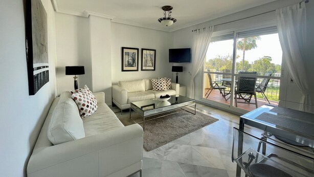 Una sala moderna con sofá, mesa de centro y televisor. Cuenta con un balcón y mobiliario exterior.