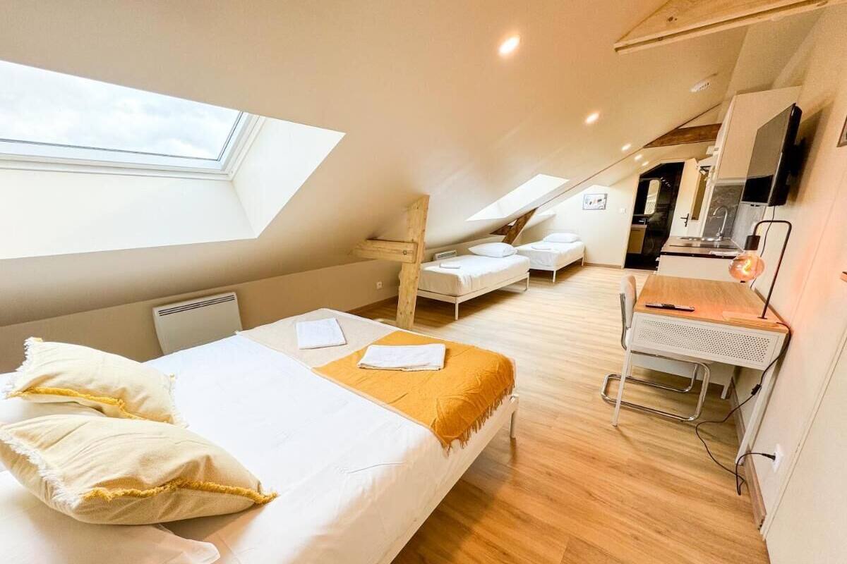 A bedroom with a skylight, a bed with pillows, a desk with a chair, and a kitchenette in the background.