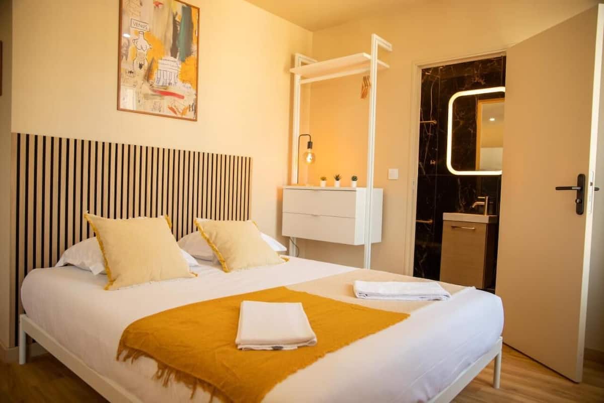 A hotel room with a bed, a bedside table with a lamp, a mirror, and a door to the bathroom.