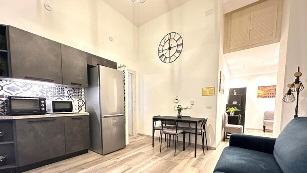 A modern kitchen with a built-in oven, microwave, and refrigerator. There is a dining area with a table and chairs, and a living room with a sofa.
