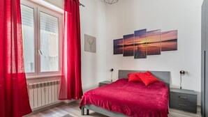 A bedroom with a bed, a nightstand, a wall-mounted lamp, and a window with curtains.