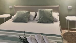 A neatly made bed with green and white pillows, a striped blanket, and a towel bundle on the side. There are two bedside tables and a wall-mounted light fixture. - Lucky Corner - Elegant Stay (Taranto)