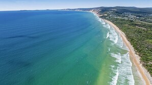 An aerial view of a coastal area with a beach, clear blue waters, and a hilly landscape in the background.