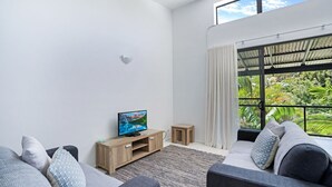 A modern living room with a flat-screen TV on a wooden stand, a sofa, a small wooden side table, and a large window with a view of greenery.