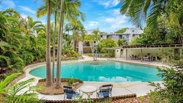 A swimming pool surrounded by palm trees and greenery, with lounge chairs and a covered seating area.