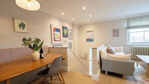 A modern living room with a wooden dining table, a sofa, and a small table with a lamp. There are framed pictures on the walls and a window with a view. - Light filled Apartment - newly renovated (London)