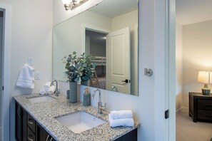 Une salle de bain avec un lavabo doté d’un plan de travail en granit, un grand miroir, une plante en pot et une table de chevet avec une lampe.