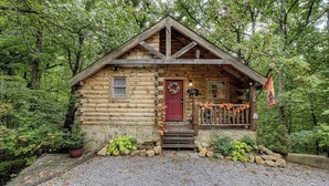 Une cabane en rondins avec une porte rouge, un petit porche doté d’une balustrade et des fondations en pierre.