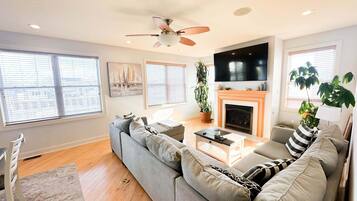 A modern living room with a gray sofa, a fireplace, a ceiling fan, and a large TV.