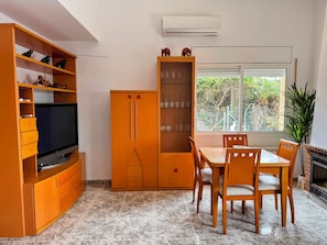 A room with a TV on a wooden cabinet, a tall cabinet with glass doors, a dining table with chairs, and a window with a view of greenery.