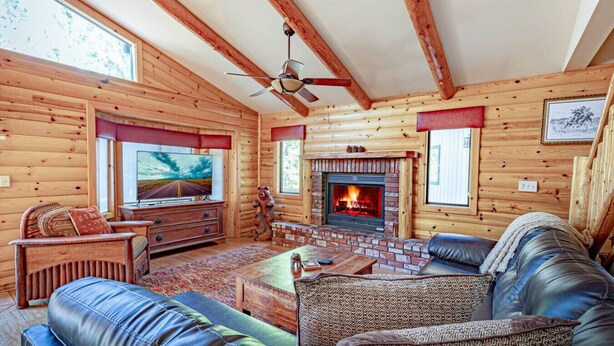 Interior de una cabaña de troncos con chimenea, sofá de cuero, paredes de madera y un televisor.